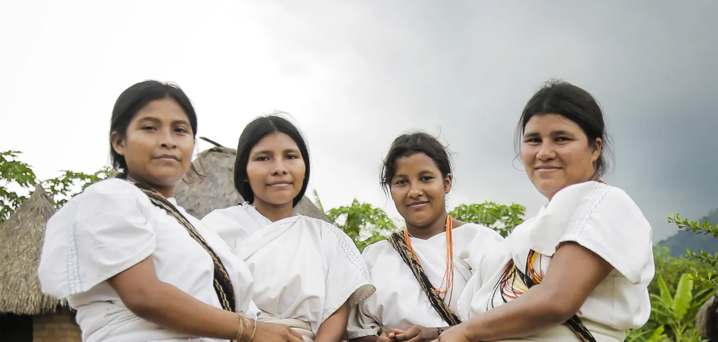 Cuatro mujeres indígenas luciendo su vestimenta tradicional con mantas de color blanco, collares y mochilas terciadas. De fondo una maloca indígena y naturaleza