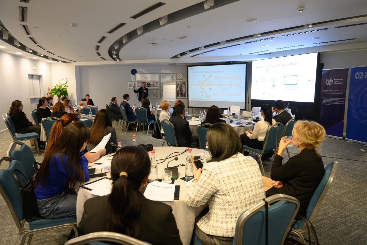 Participants of the workshop on the design and implementation of PWPs sit at round tables and listen to the presenter in front of the screen on 6 February 2025 in Armenia