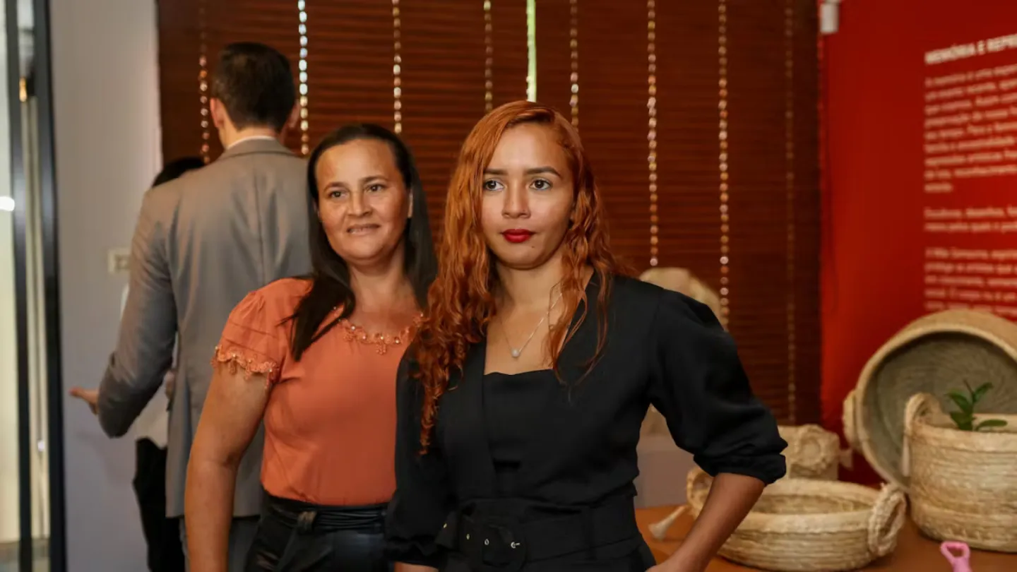 Fátima Laine Sousa Santos está em frente a uma exposição de produtos artesanais de carnaúba trançados em um evento indoor. Outra participante do evento está ao lado dela e sorri.