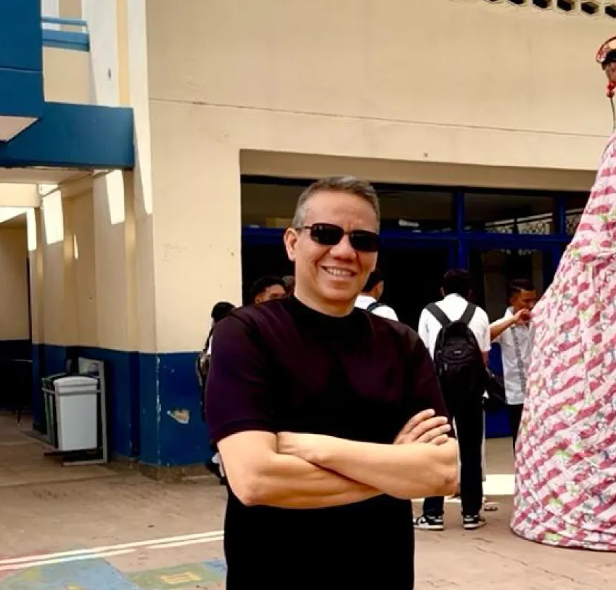 Hombre cruzado de brazos con camiseta y gafas negras al frente de un edificio, detrás de él se ven jóvenes uniformados con maletas