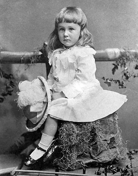 Fotografía en blanco y negro de un niño de cabello láceo, largo, luciendo un vestido y zapatos de charol, sosteniendo un sombrero con arreglos, sentado sobre la imitación de un tronco en un set fotográfico en el que en el fonde se ha puesto una cerca de madera con enredaderas de plantas artificiales