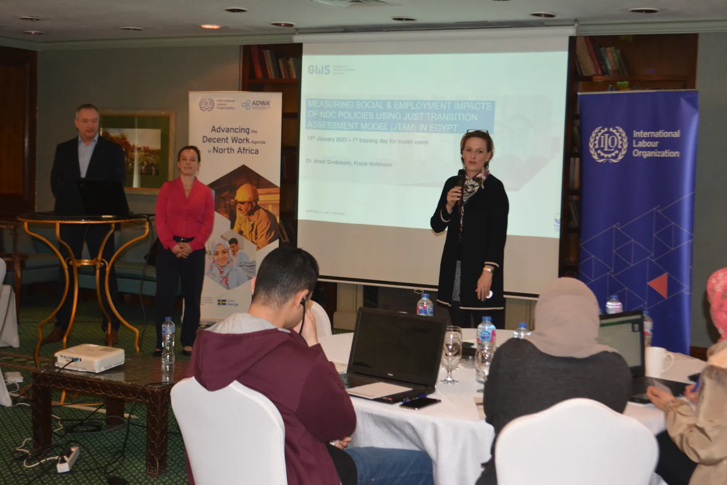 A woman speaks into a microphone during a training workshop, while two facilitators stand near a presentation screen displaying a slide. Participants are seated at round tables with laptops.