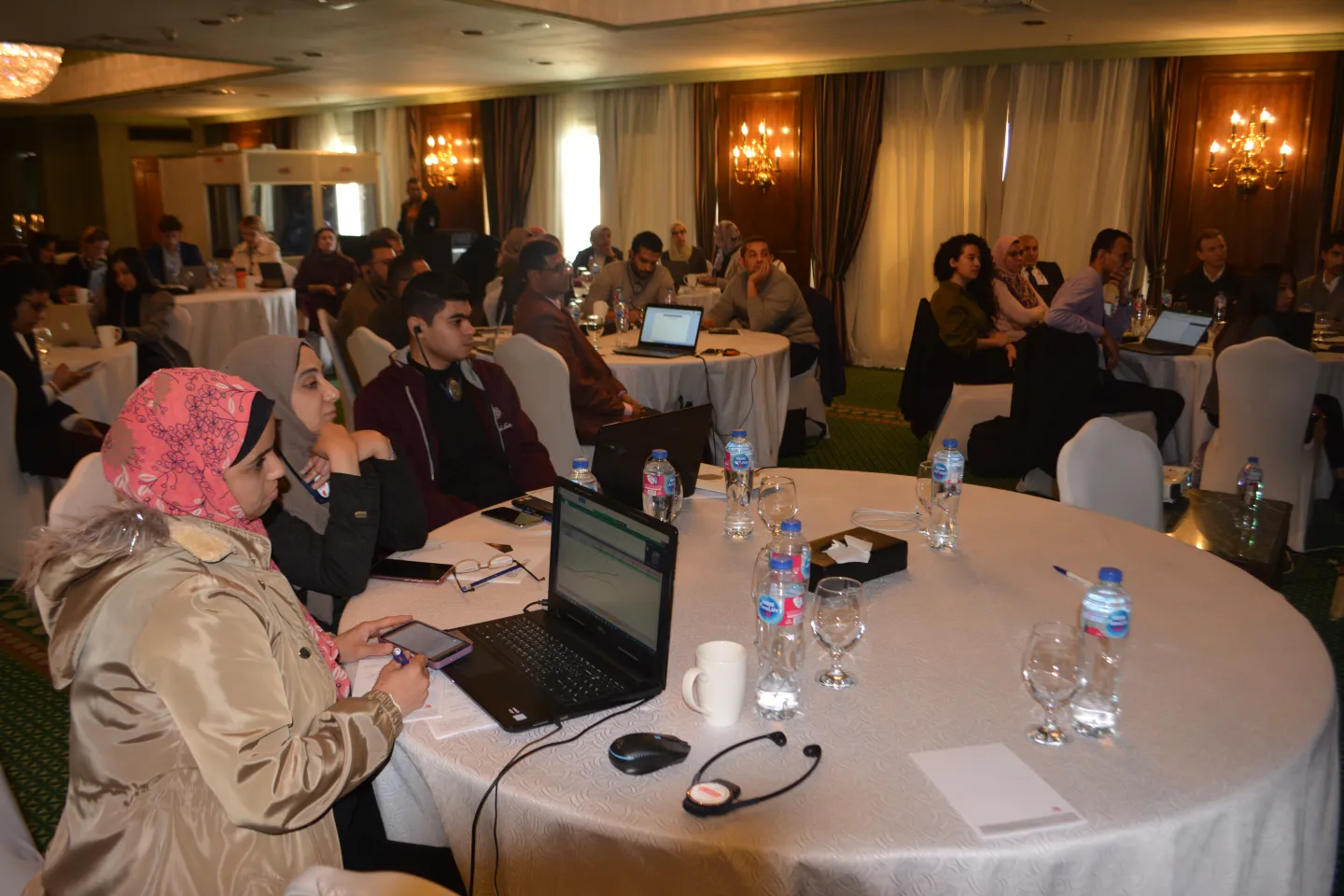 Groups of participants seated at round tables with laptops and documents during a training workshop in a hotel conference room, listening attentively.