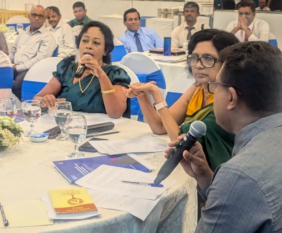 A lady in a sari, seated at a table, addresses a room full of participants