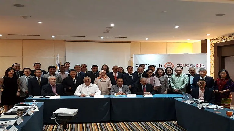 A group of persons sitting and standing in a meeting room. In the background, a banner with logos from ACE, ATUC, the ILO, and DFAT and GAC are partially visible.