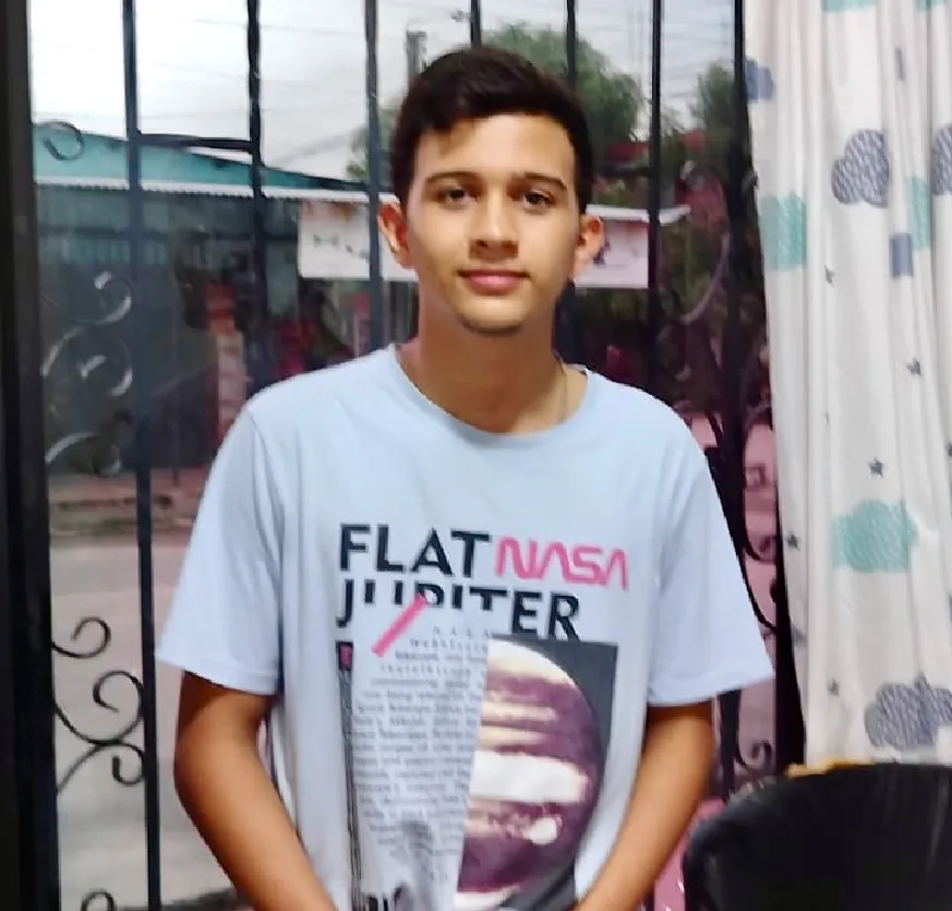 Adolescente de 16 años, está de pie en el interior de su casa. Tiene el cabello corto y oscuro, la piel clara y expresión serena. Lleva puesta una camiseta celeste clara. A su alrededor se ven detalles del hogar: cortinas blancas decoradas con nubes azules y grises, una silla negra de oficina, una mesa con cuadernos y objetos escolares