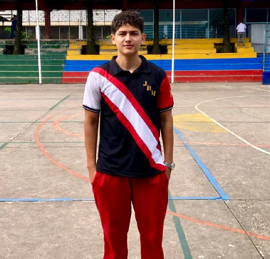 Joven de 16 años, está de pie en el centro de una cancha polideportiva al aire libre. Viste el uniforme de su institución educativa: una camiseta tipo polo negra con detalles en blanco y rojo en diagonal, con las letras "JBM", siglas de Juan Bautista Migani, bordadas en el pecho, y pantalones deportivos rojos.