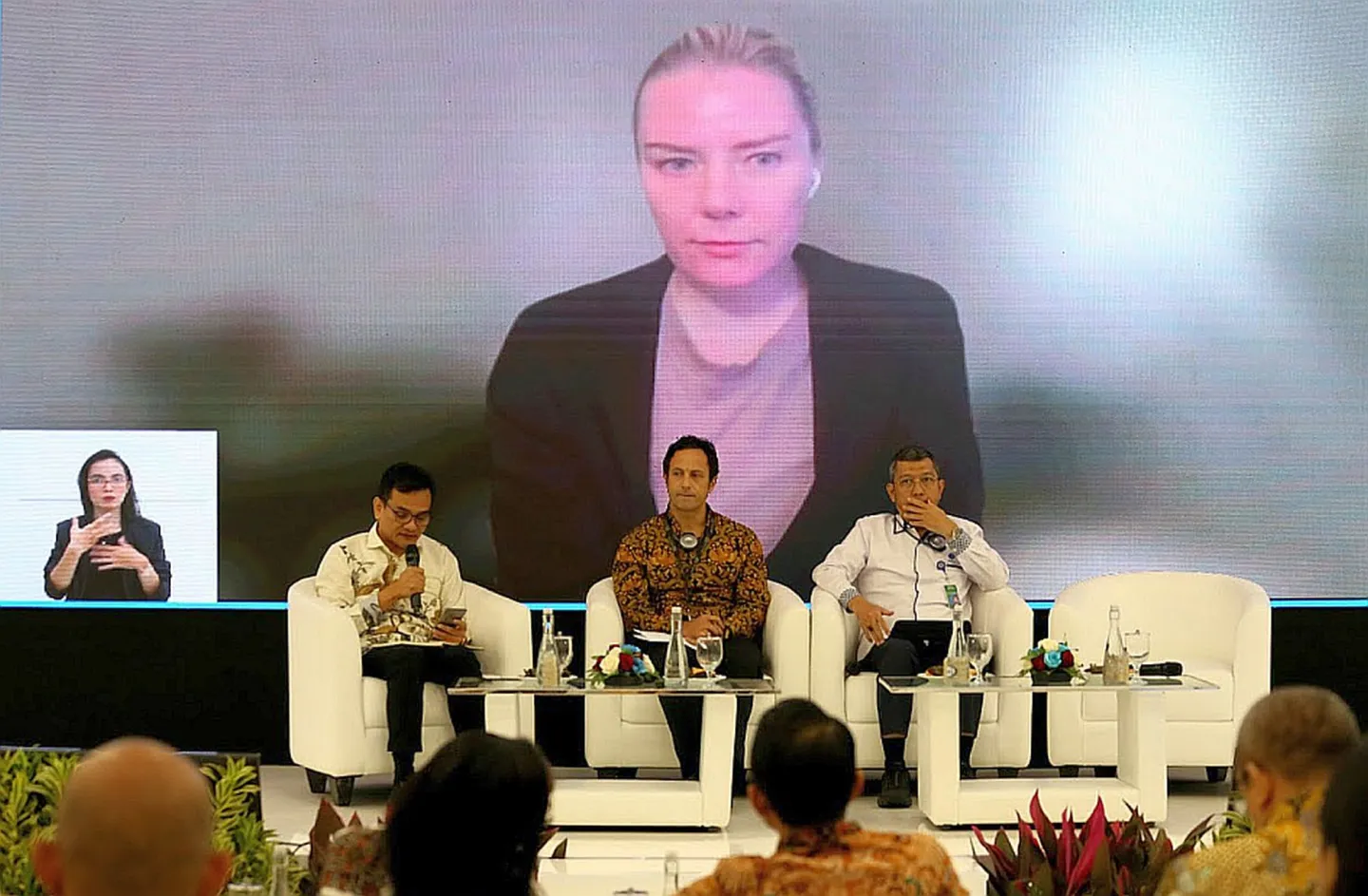 Three persons sitting on stage with a screen on the back featuring another speaker