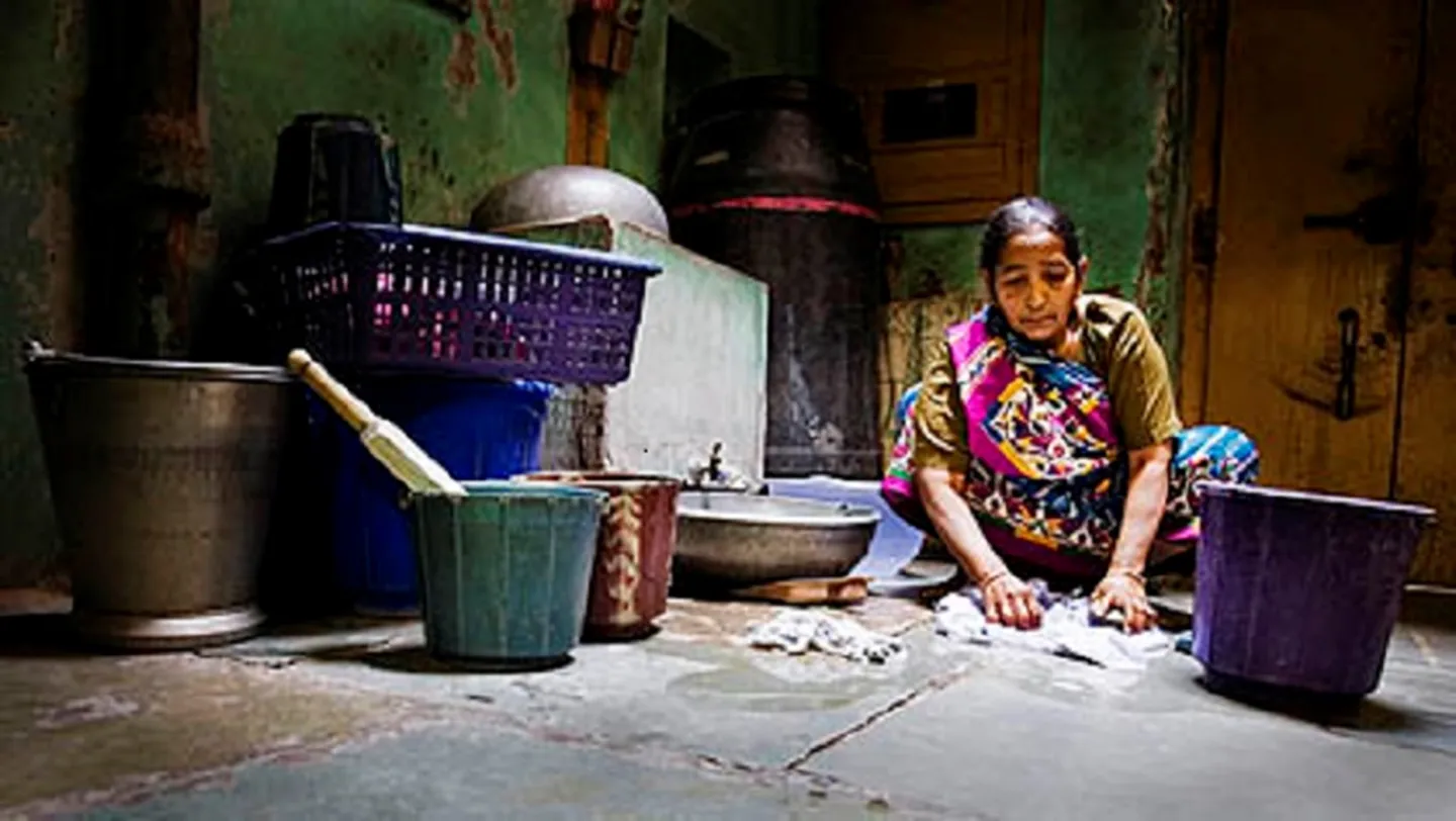 A person is washing clothes by hand in a dimly lit room surrounded by several buckets and containers.
