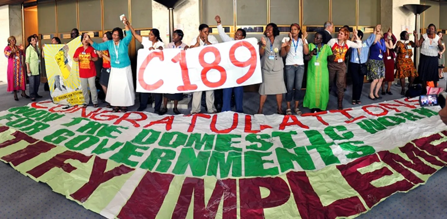 A group of people stands behind a large banner reading "CONGRATULATIONS FOR THE DOMESTIC WORK GOVERNMENTS RATIFY IMPLEMENT" and holds a smaller banner labeled "C189."
