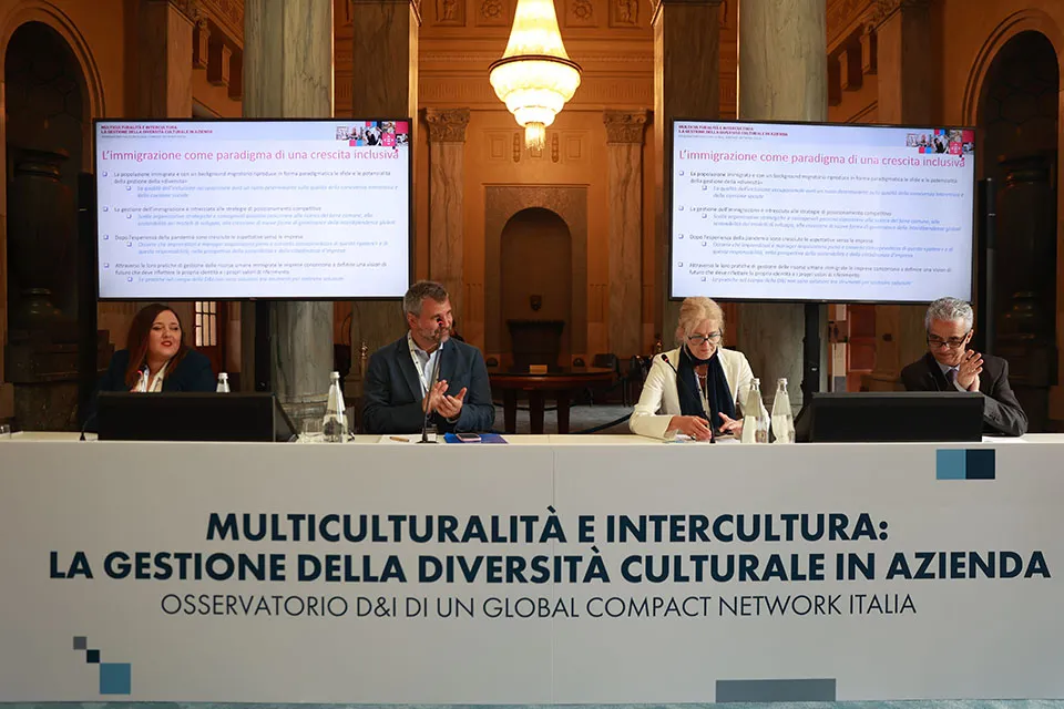 Quattro persone sedute al tavole dei relatori di una conferenza, con due megaschermi alle spalle.