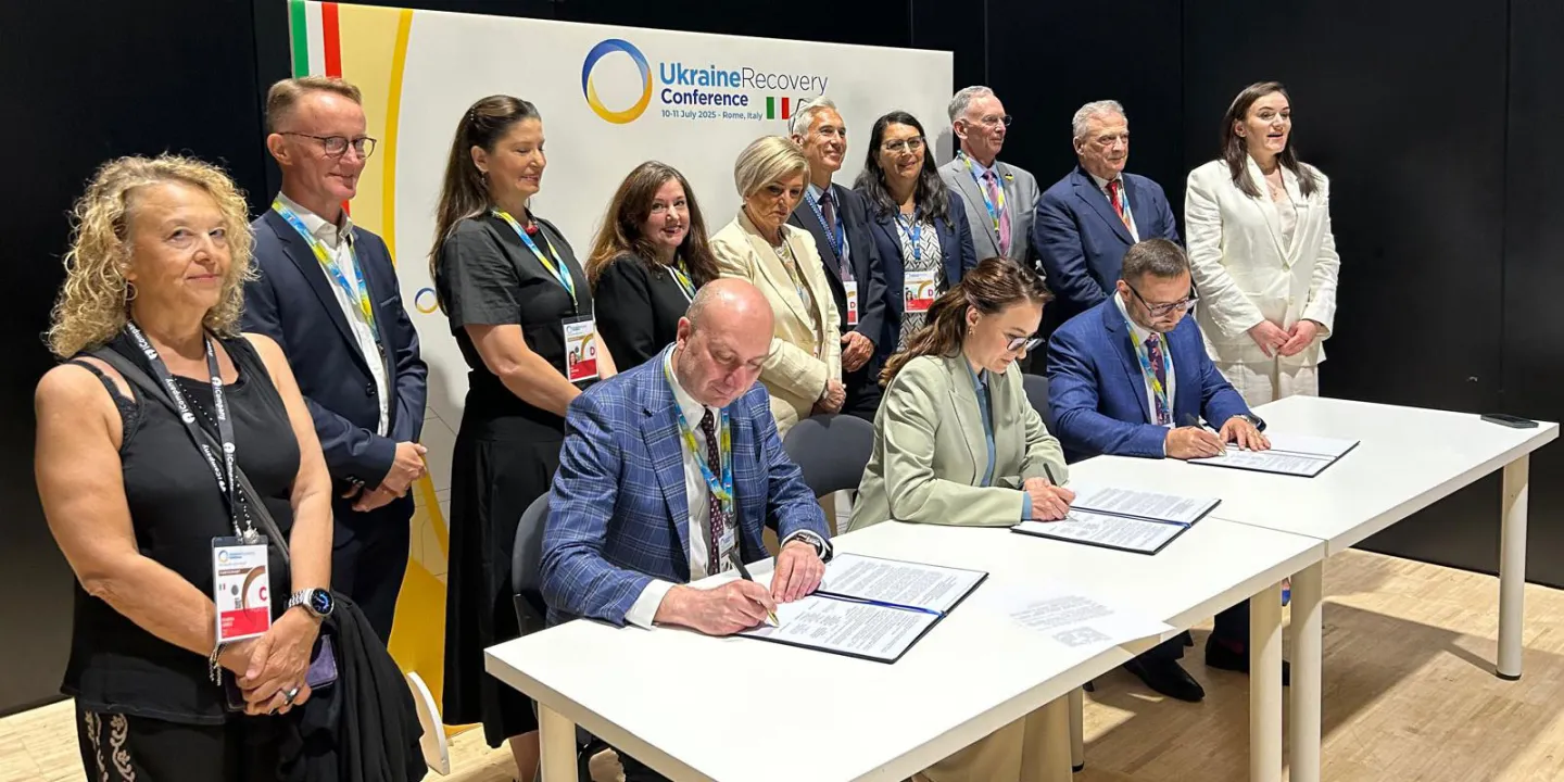Members of official delegations surrounding a table with three persons who are signing an agreement