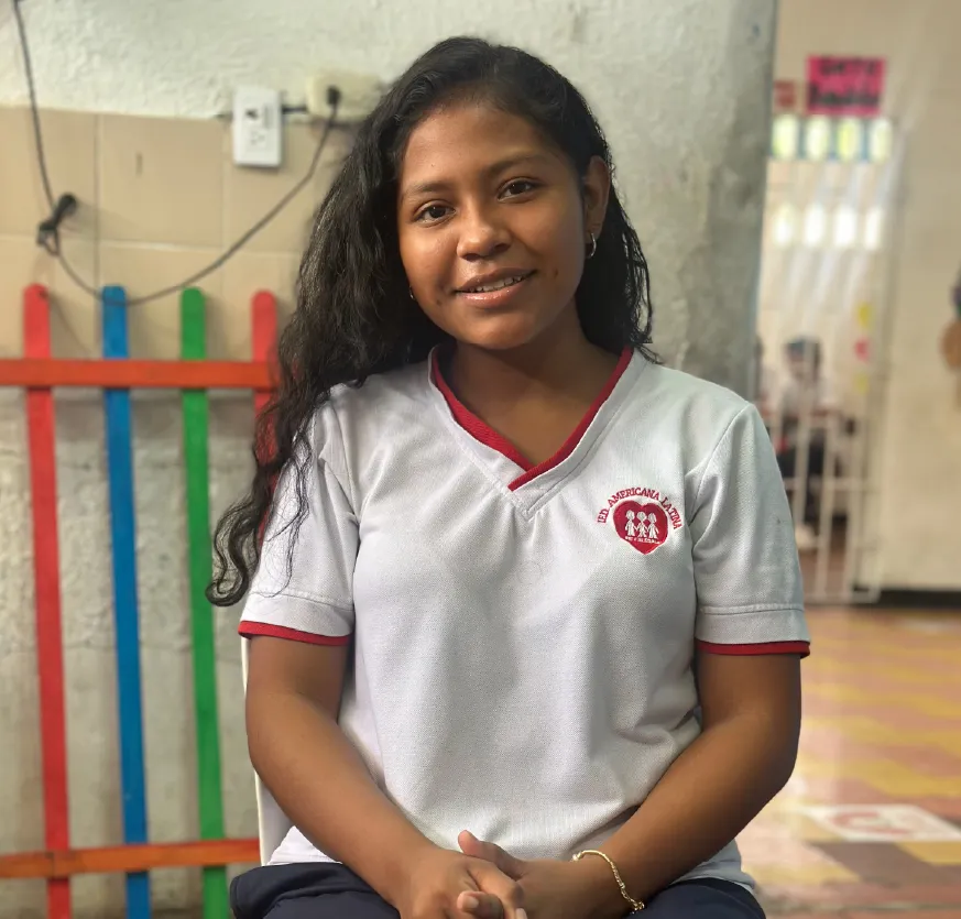 Joven mujer de tes morena, cabello negro y largo, mira de frente a la cámara y sonríe; está sentada en una silla, tiene las manos entrelazadas sobre las piernas. Viste un pantalón azul oscuro y una camiseta de mangas cortas de color blanco. Al costado izquierdo del pecho tiene estampado un corazón rojo logotipo de la organización Fe y Alegría, en la parte superior del corazón rojo hay un texto con el nombre de la Institución Educativa Distrital América Latina. De fondo se ve el colegio donde estudia. 