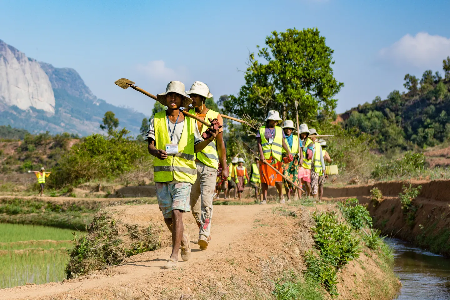 Workers in protective gear carrying tools walking along an irrigation ditch