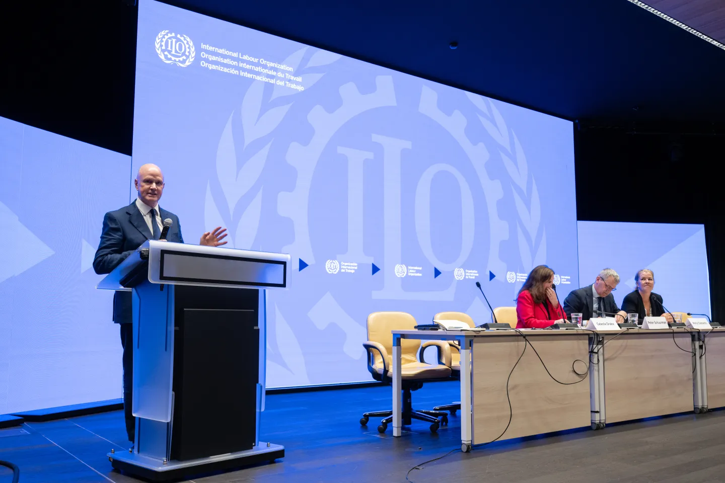 person speaking at the podium while panel members listen to him
