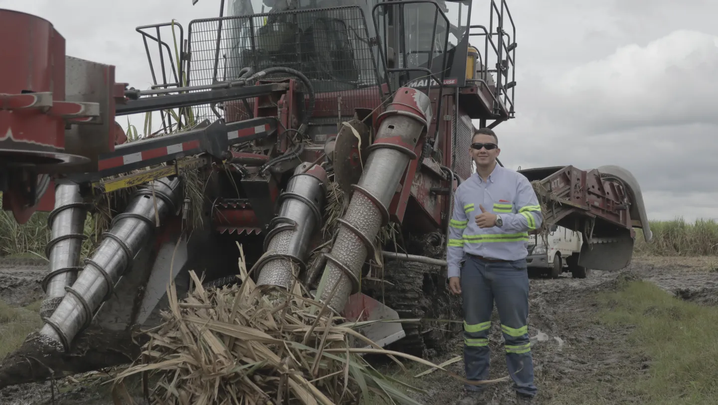 Hombre con gafas oscuras y camisa de trabajo con franjas reflectantes de pie sobre un terreno fangoso, al lado de una cosechadora de caña de azúcar de gran tamaño.