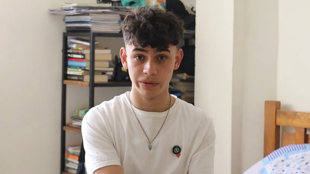  Un joven de 16 años, aparece sentado en su habitación. Tiene la piel clara y el cabello oscuro y rizado peinado con volumen en la parte superior. Viste una camiseta blanca y lleva en el cuello una cadenilla con un dije triangular. Su expresión es seria y atenta, con la mirada directa hacia la cámara. Detrás de él se ve una estantería metálica alta, repleta de libros, cuadernos y papeles apilados. 