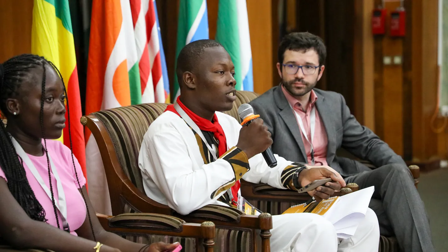 Youth delegate speaks into a microphone during a panel discussion at the 4th Annual West Africa and Sahel Youth Forum in Dakar, seated alongside fellow participants with country flags in the background.