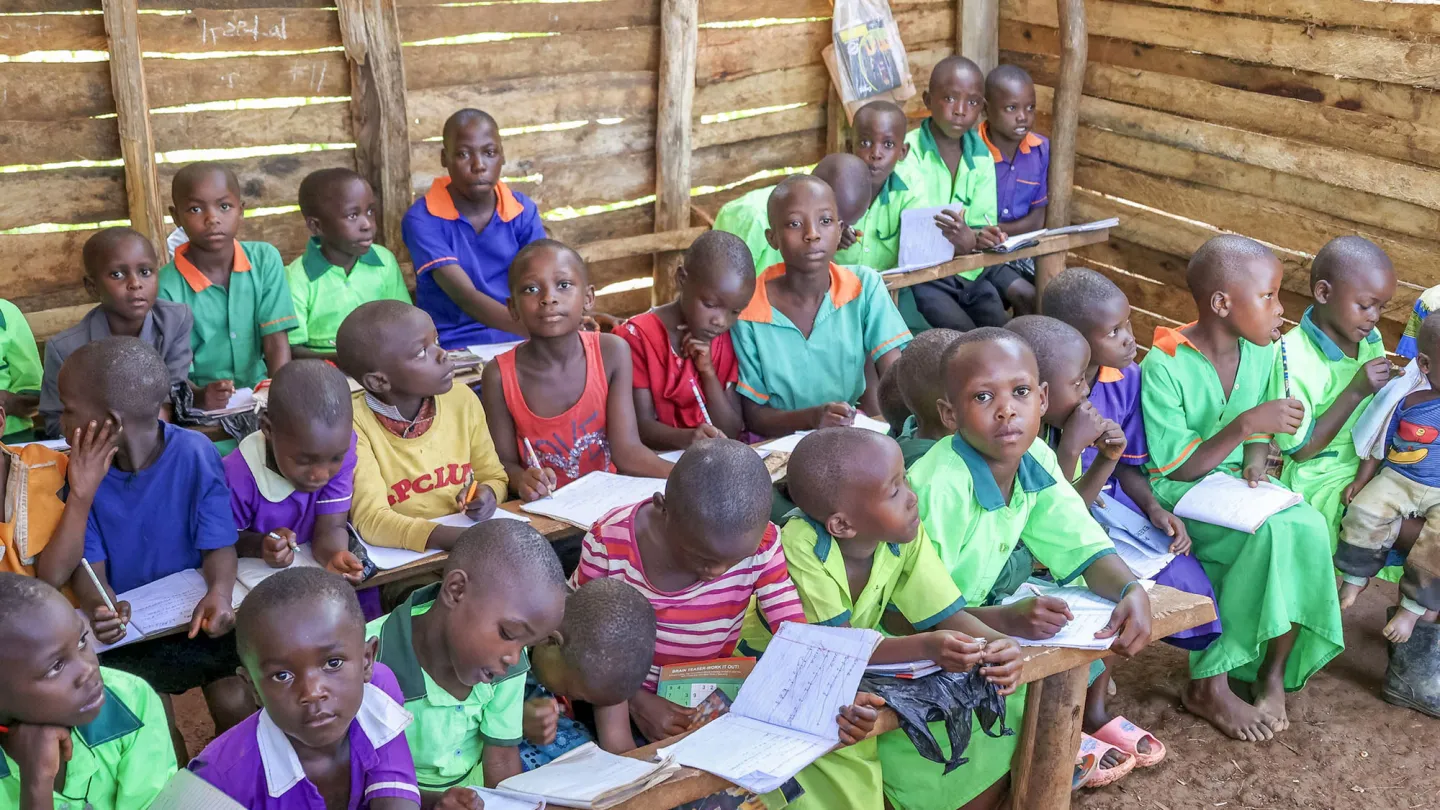 A classroom of young children attentively writing in their notebooks inside a wooden school building in rural eastern Uganda.