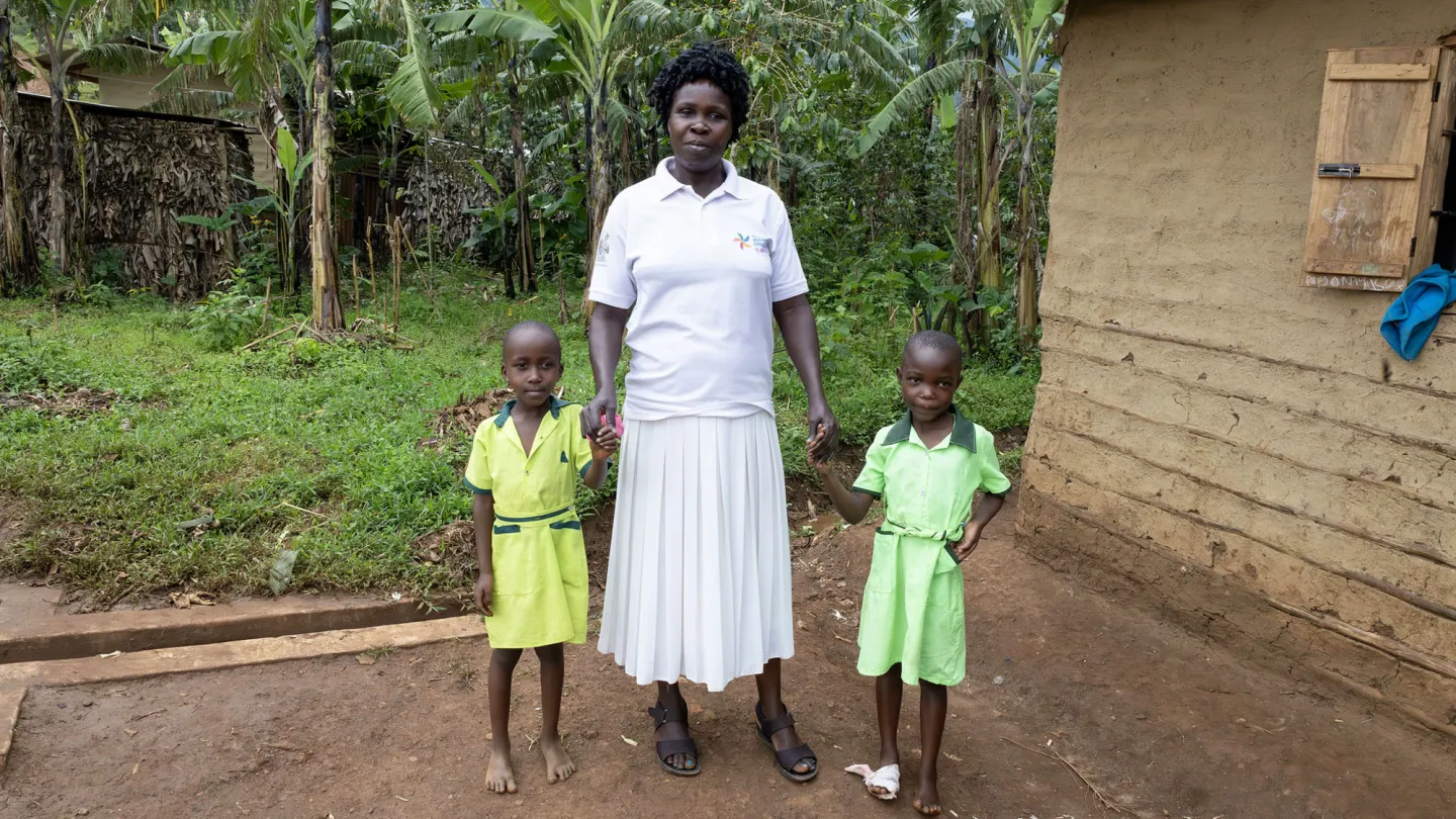A woman stands holding hands with her two young children outside a school building in rural eastern Uganda, surrounded by green vegetation.