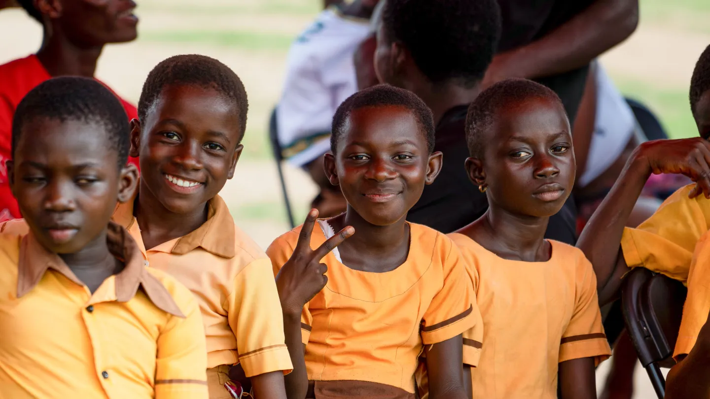A group of children in yellow school uniforms sit and smile at the camera during a visit by the Japanese Ambassador to their school in Beposo, Ghana. One child flashes a peace sign, while others look on. The visit was part of the Climate Cocoa Project's support to child labour-free zones.
