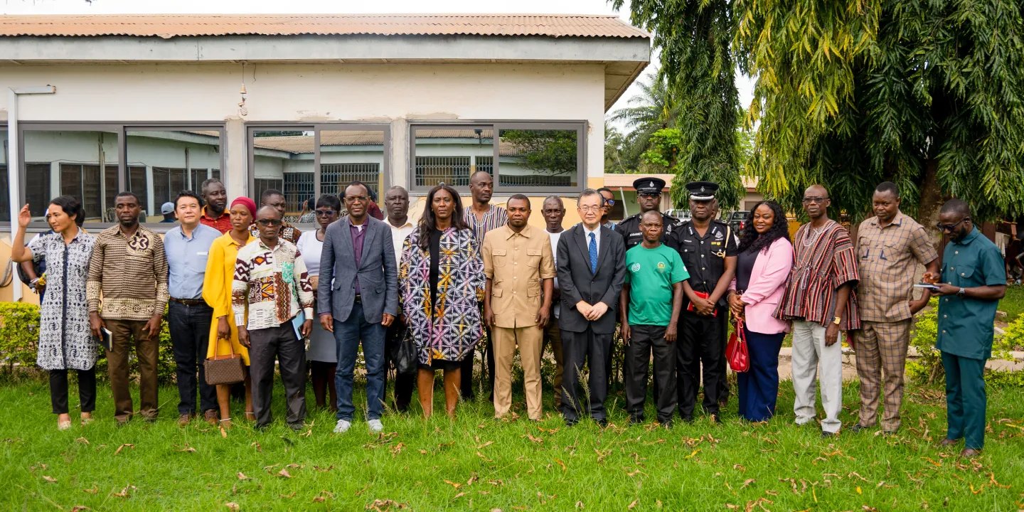 Group photo of the Japanese Ambassador to Ghana, ILO officials including Country Director Vanessa Phala and Deputy Regional Director Coffi Agossou, standing with municipal authorities, police officers, and community members in front of a building in Bibiani, Ghana. The photo was taken during a high-level mission to promote child labour-free zones under the Climate Cocoa Project.