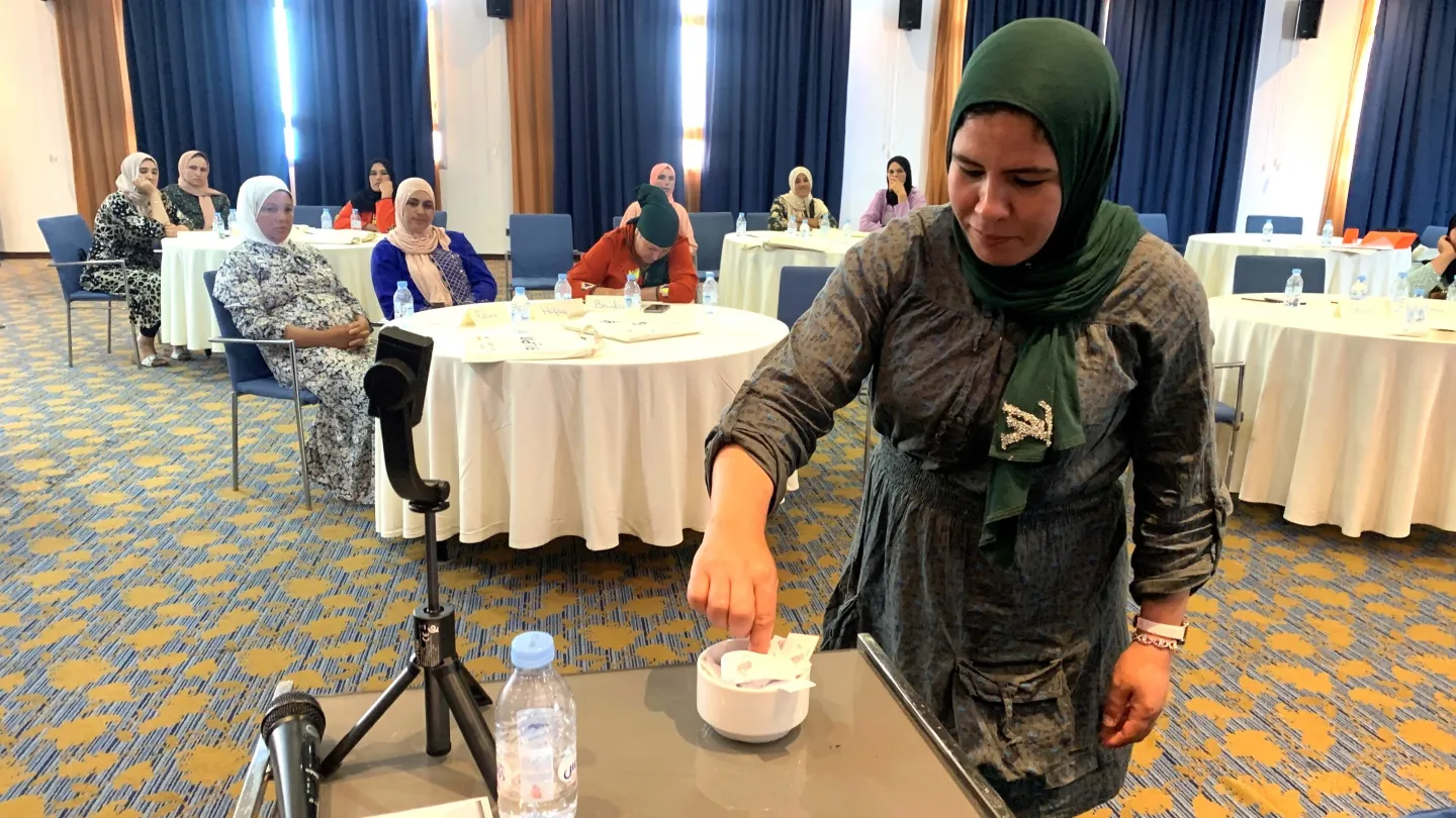 Fatima places a piece of paper in a dish in the foreground of the image. Fatima and other women are in a large room with round tables where a WAFIRA training session is taking place. 