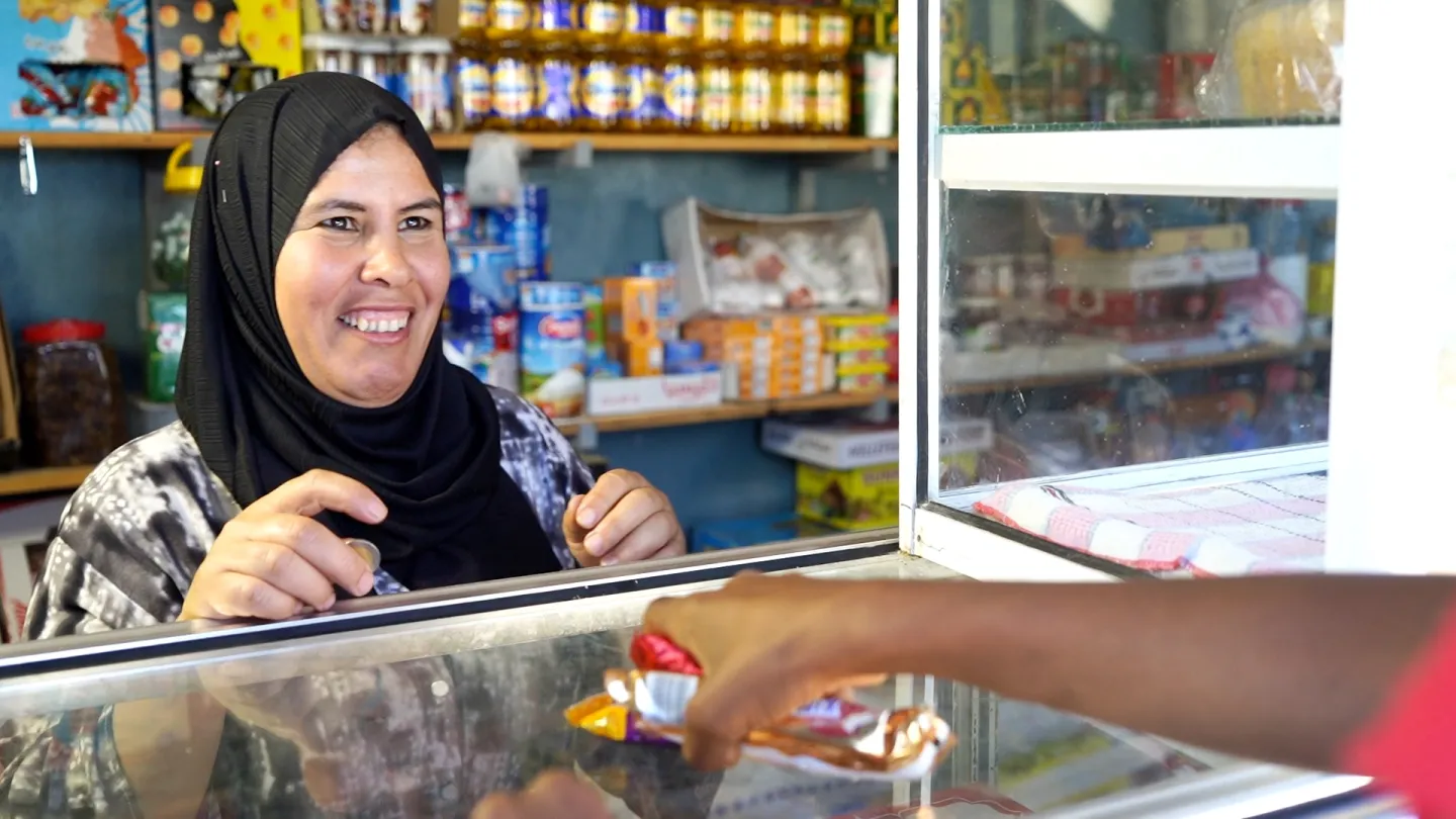 Fatima stands at the counter of her shop and smiles at her customer.  The customer’s hand is taking some sweets they have bought, and Fatima holds the payment, a coin, in her hand.
