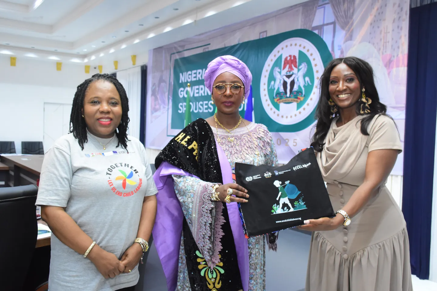 Three people stand indoors at an event. One person in traditional attire holds a black bag with printed logos while another person hands it over.