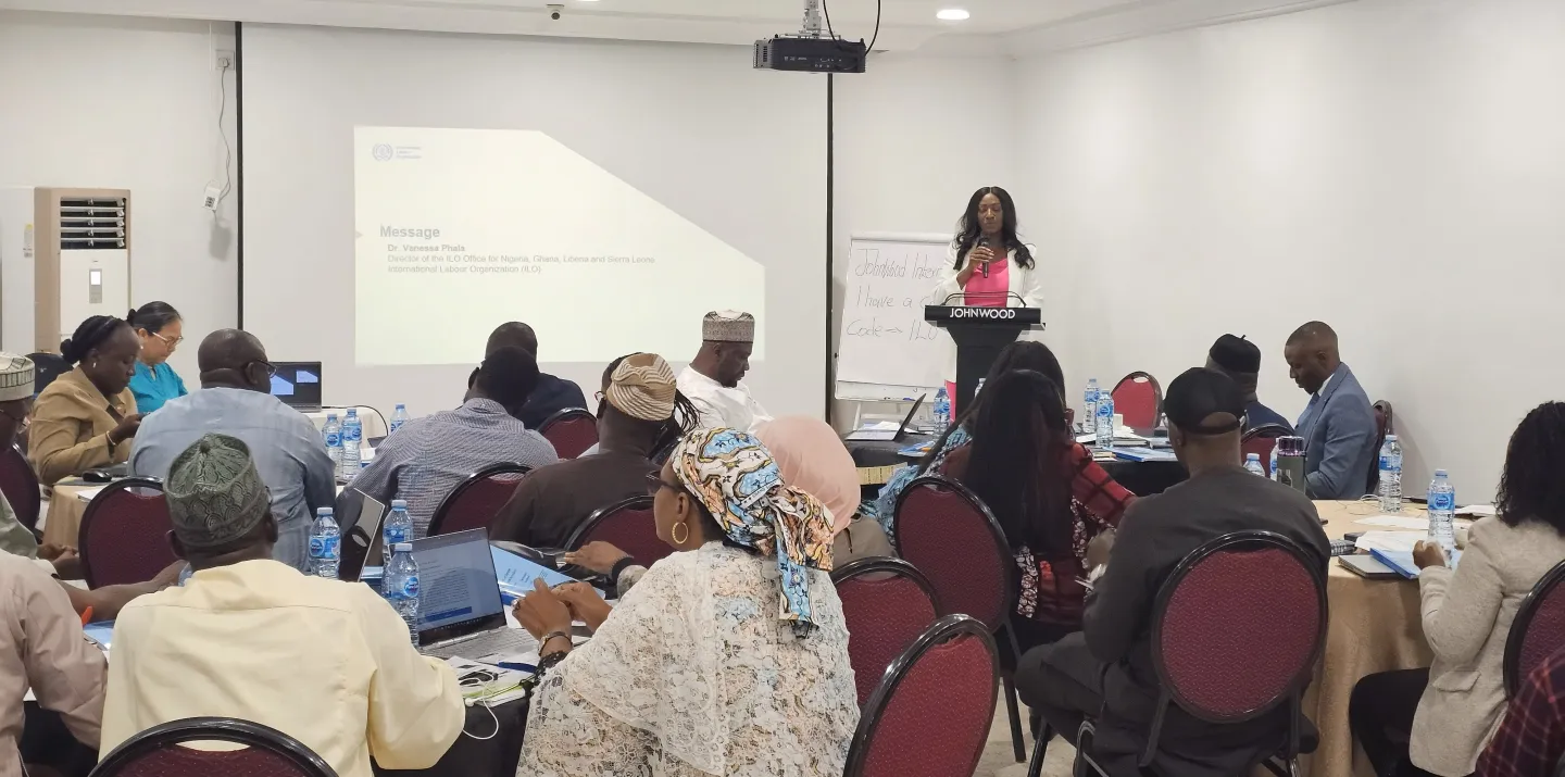 A speaker stands at a podium addressing participants seated around tables during a workshop.