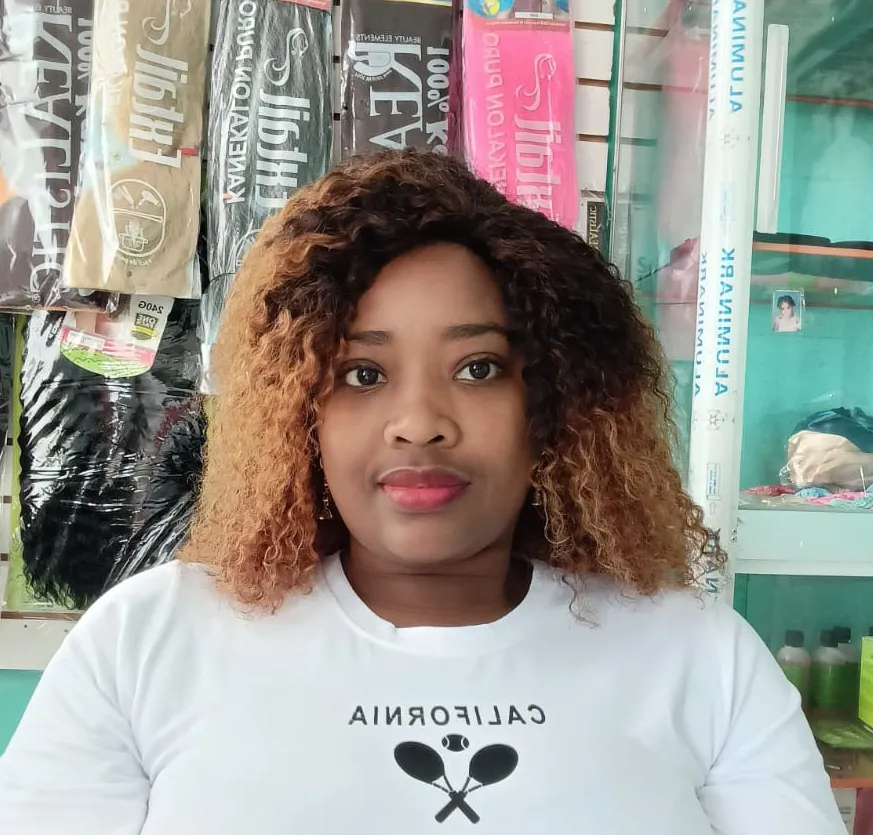 La imagen muestra a una mujer joven sentada dentro de una tienda de productos de belleza. Ella tiene el cabello rizado, con raíces oscuras y puntas color miel. Su expresión es seria y tranquila. Lleva una camiseta blanca con letras grandes en el pecho. Detrás de ella se ven estantes con extensiones de cabello empacadas y otros productos de estética organizados en vitrinas de vidrio. El ambiente es luminoso y ordenado.