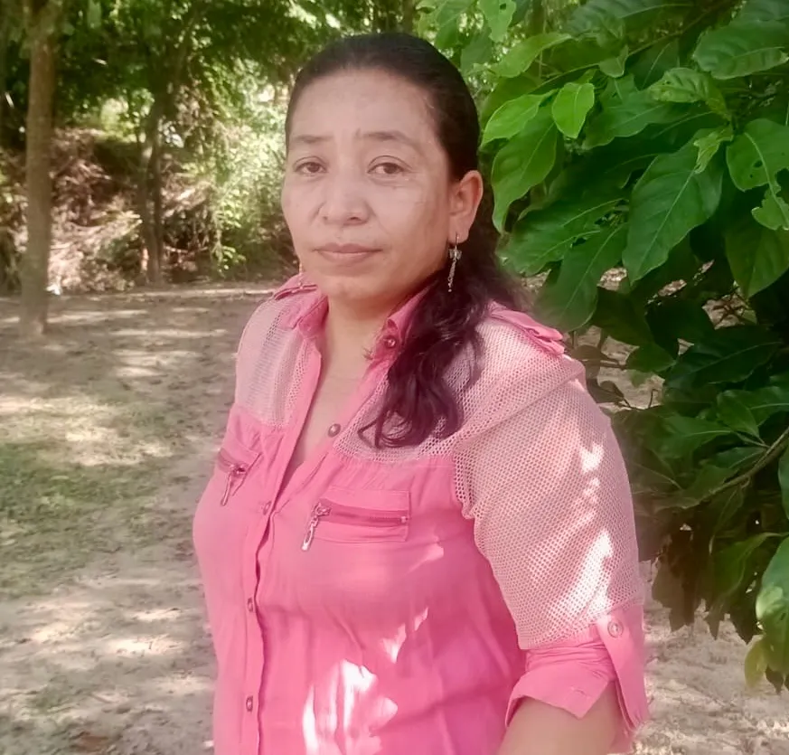 La foto muestra a una joven mujer de piel morena sentada frente a una mesa. Lleva puesta una camiseta tipo polo blanca con los logos del Ministerio del Trabajo y la Organización Internacional del Trabajo (OIT) en el pecho. Tiene el cabello recogido en trenzas y mantiene los brazos cruzados sobre la mesa. Mira de frente a la cámara con una expresión tranquila y segura. El fondo es claro y sencillo, con una cortina blanca a un lado.