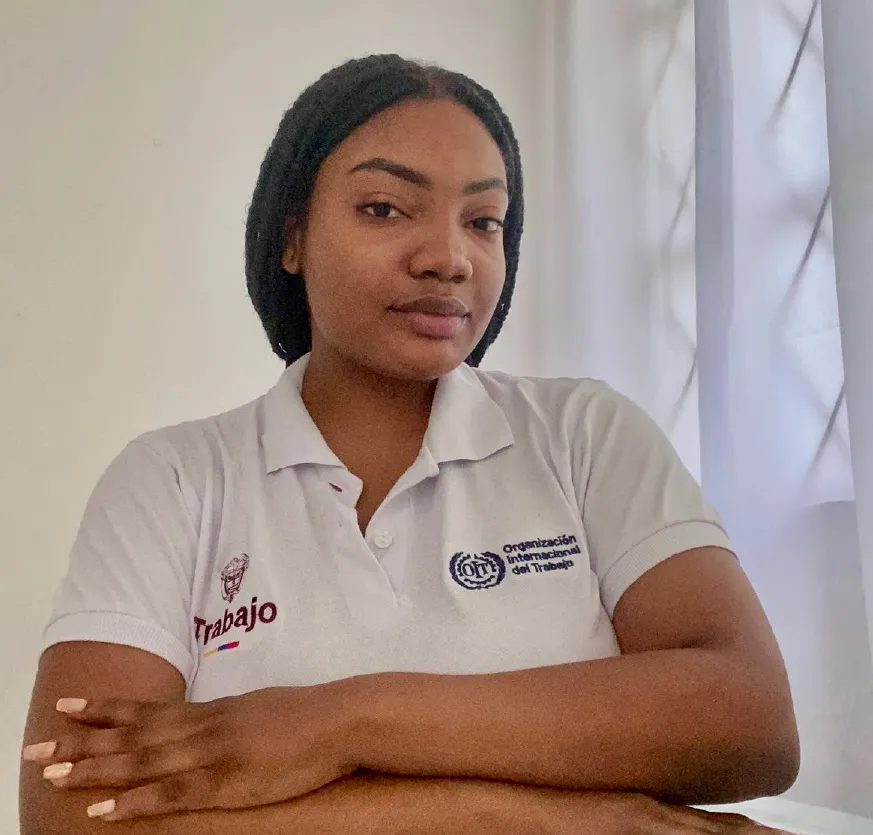 La foto muestra a una joven mujer de piel morena sentada frente a una mesa. Lleva puesta una camiseta tipo polo blanca con los logos del Ministerio del Trabajo y la Organización Internacional del Trabajo (OIT) en el pecho. Tiene el cabello recogido en trenzas y mantiene los brazos cruzados sobre la mesa. Mira de frente a la cámara con una expresión tranquila y segura. El fondo es claro y sencillo, con una cortina blanca a un lado.