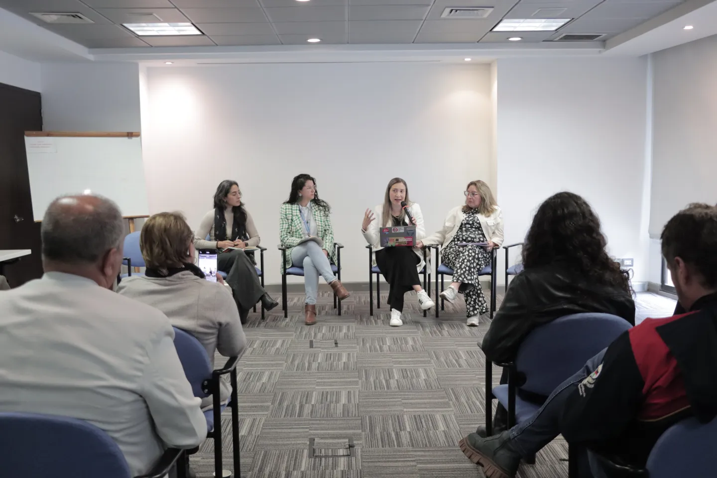 En esta imagen se observa un panel de conversación en una sala de reuniones. Cuatro personas están sentadas al frente, formando una fila, participando en un diálogo frente a un grupo de asistentes. Una de ellas sostiene un micrófono y habla, mientras las demás escuchan con atención. Frente a ellas, el público permanece sentado en sillas azules, observando o grabando con un teléfono móvil.
