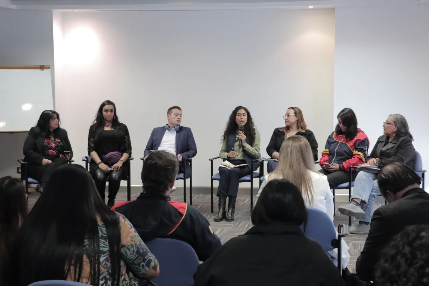 Se observa un panel de conversación en un auditorio. Siete personas están sentadas al frente, formando una fila, mientras dialogan frente a un público. Una de ellas sostiene un micrófono y parece estar interviniendo. Las demás escuchan con atención, algunas con cuadernos o teléfonos en las manos. En primer plano se ven los asistentes sentados en filas de sillas azules.