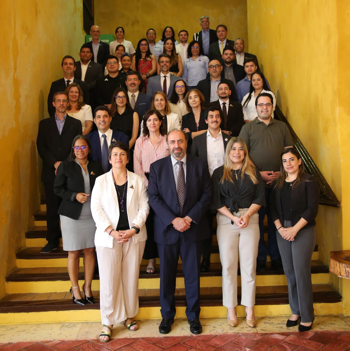 Grupo de aproximadamente treinta personas posando en una escalera interior de paredes amarillas. Están organizadas en varias filas, la mayoría vestidas con ropa formal y de oficina, algunas sonriendo hacia la cámara.