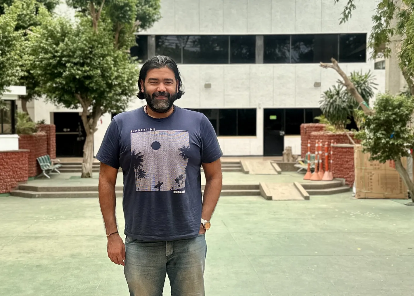 Un hombre está de pie en un patio al aire libre. Tiene cabello oscuro y largo hasta los hombros, peinado hacia atrás, y barba completa. Viste camiseta azul con un estampado de palmeras y un surfista, y lleva jean azul claro. Sonríe levemente, con la mano derecha en el bolsillo y la izquierda relajada al costado. Detrás de él hay árboles, bancas verdes y un edificio blanco de dos pisos con ventanas negras. A la derecha se observan conos naranjas de seguridad agrupados.