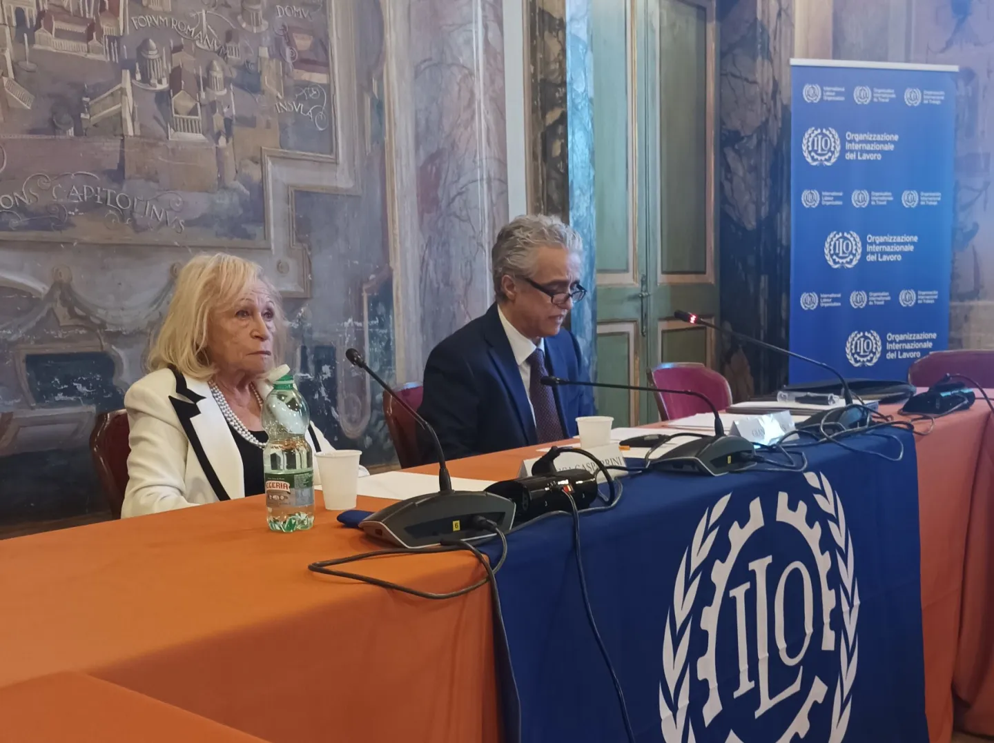 Una donna e un uomo seduti al tavolo dei relatori di una sala conferenza.