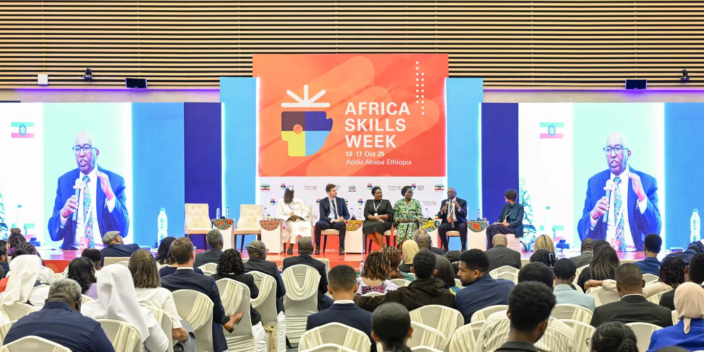 A panel of six speakers sits on stage at Africa Skills Week 2025 in Addis Ababa, addressing a seated audience. A large screen displays a close-up of one speaker holding a microphone.