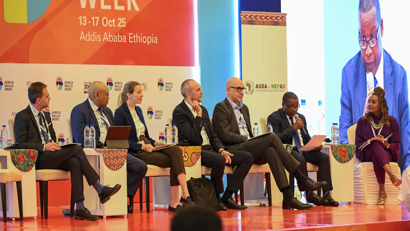 A diverse panel of seven speakers sits on stage during Africa Skills Week 2025 in Addis Ababa, with one speaker addressing the audience using a microphone. A large screen beside them displays a close-up of the speaker.