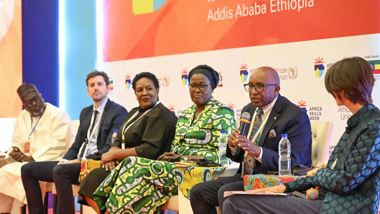 Seven panelists seated on stage at Africa Skills Week 2025 in Addis Ababa, with one speaker in a suit and glasses holding a microphone while addressing the audience.
