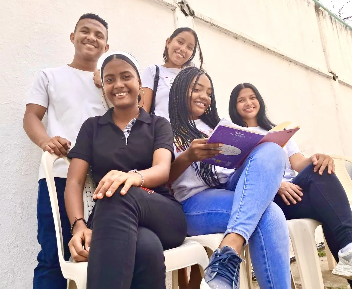 Cinco jóvenes sonríen mientras participan en una actividad de estudio al aire libre. Dos de ellas leen un cuaderno morado, mientras los demás las acompañan sentados y de pie, mostrando un ambiente de compañerismo y aprendizaje.