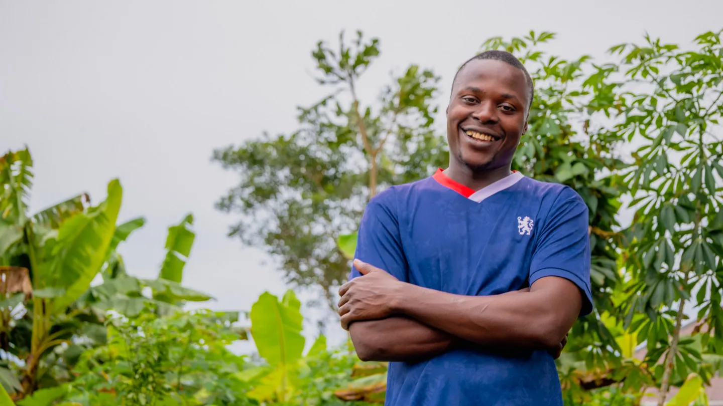 A young Nigerian man, David Chukwuemeka, stands outdoors smiling with his arms crossed, surrounded by lush green plants and trees in his rural community in Aponmu, Ondo State.