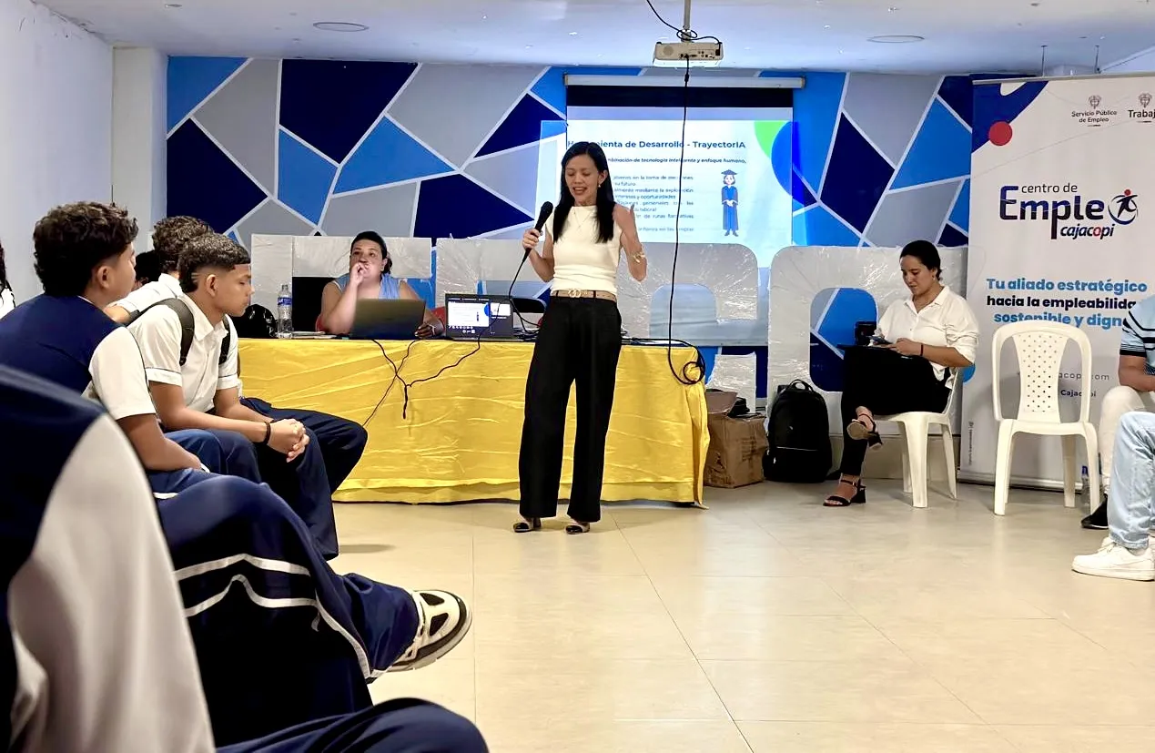 El ambiente es un salón amplio y luminoso. Una mujer de pie sostiene un micrófono y habla frente a un grupo de estudiantes sentados en círculo. Detrás de ella hay una mesa con un computador y otra mujer que escucha con atención, de fondo una pantalla que proyecta una presentación. A un costado se ve otra mujer sentada y atrás de ella un pendón del Centro de Empleo Cajacopi, en la parte superior del pendón los logos del Servicio Público de Empleo y del Ministerio del Trabajo. 