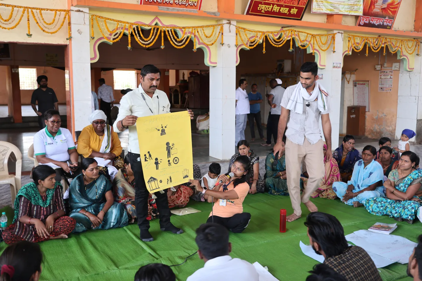 People are seated in a circle, many of them are women who wear saris.  Lata Korde sits in the middle of the circle. She holds a microphone and points at a poster that a man is holding.