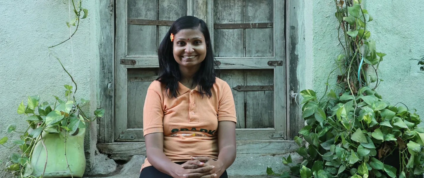 A portrait of Lata Korde. She sits on a stone step in front of a wooden door. Plants grow up a blue wall either side of the steps. (India 2024)