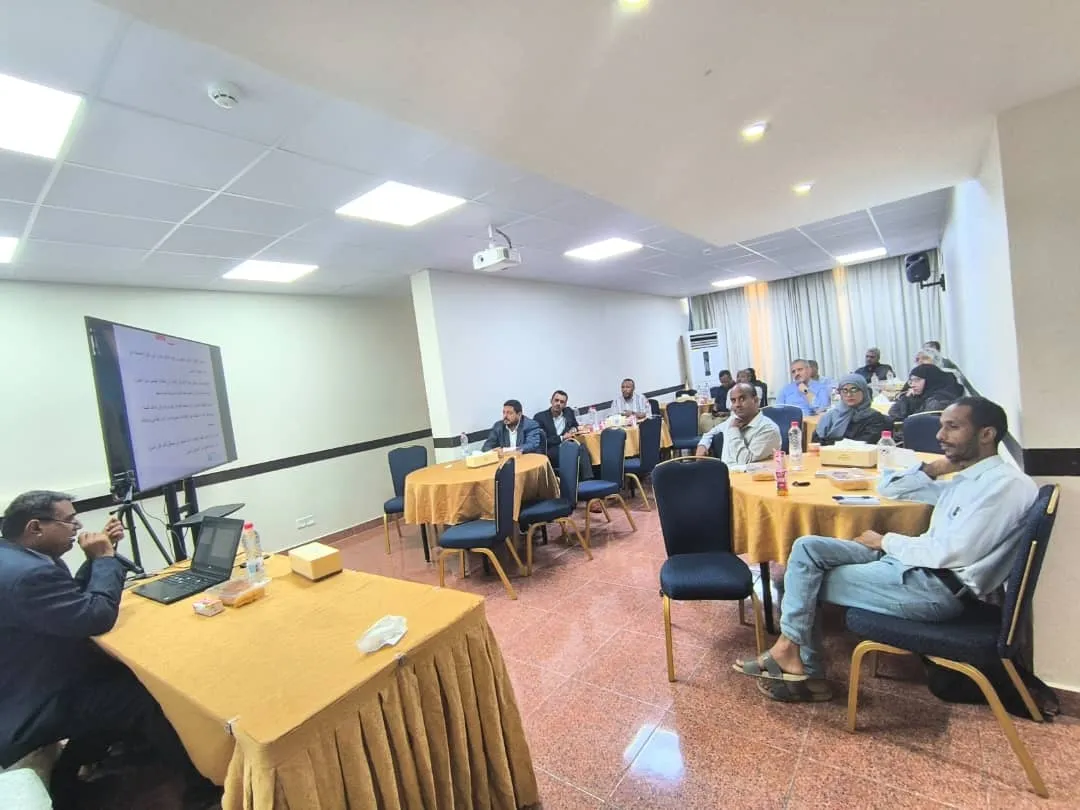 Classroom with business representatives listening to one of the main speakers. The room has a main table where the speaker is sitting, and participants are sitting in round tables.