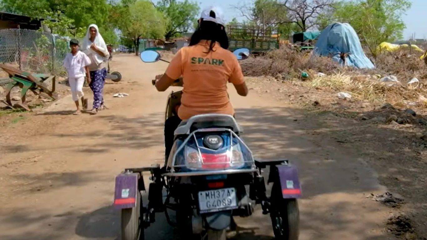 Lata Korde drives a quad bike along a dusty road. A woman and boy walk along the road in the background.