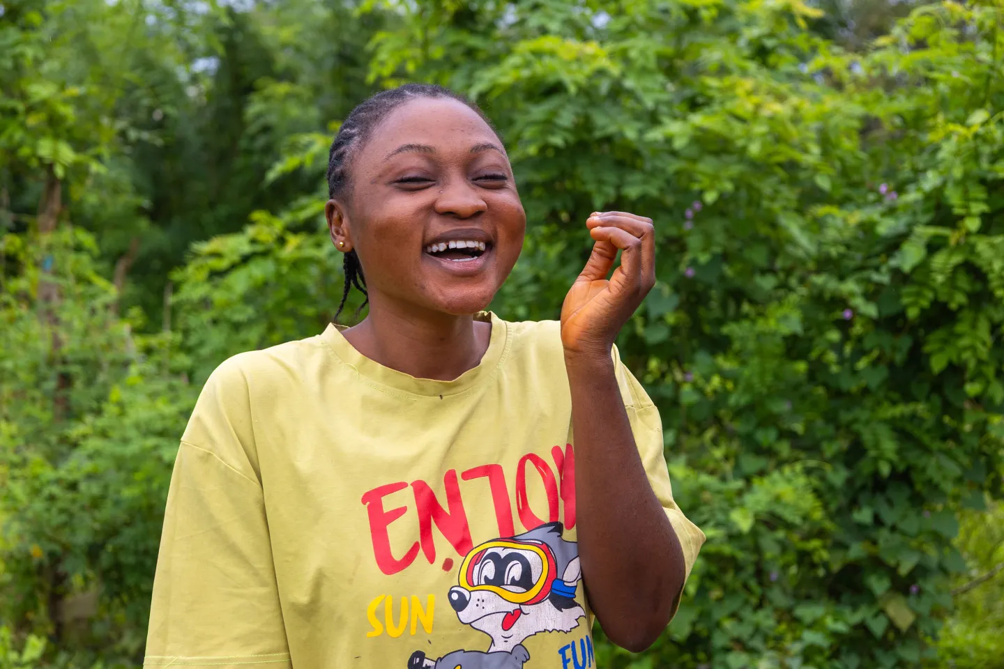 A smiling young woman, Oyefunde Bukola, laughs with her hand raised slightly as she stands outdoors in her community in Ondo State, Nigeria. Lush green foliage surrounds her, and she appears relaxed and joyful.