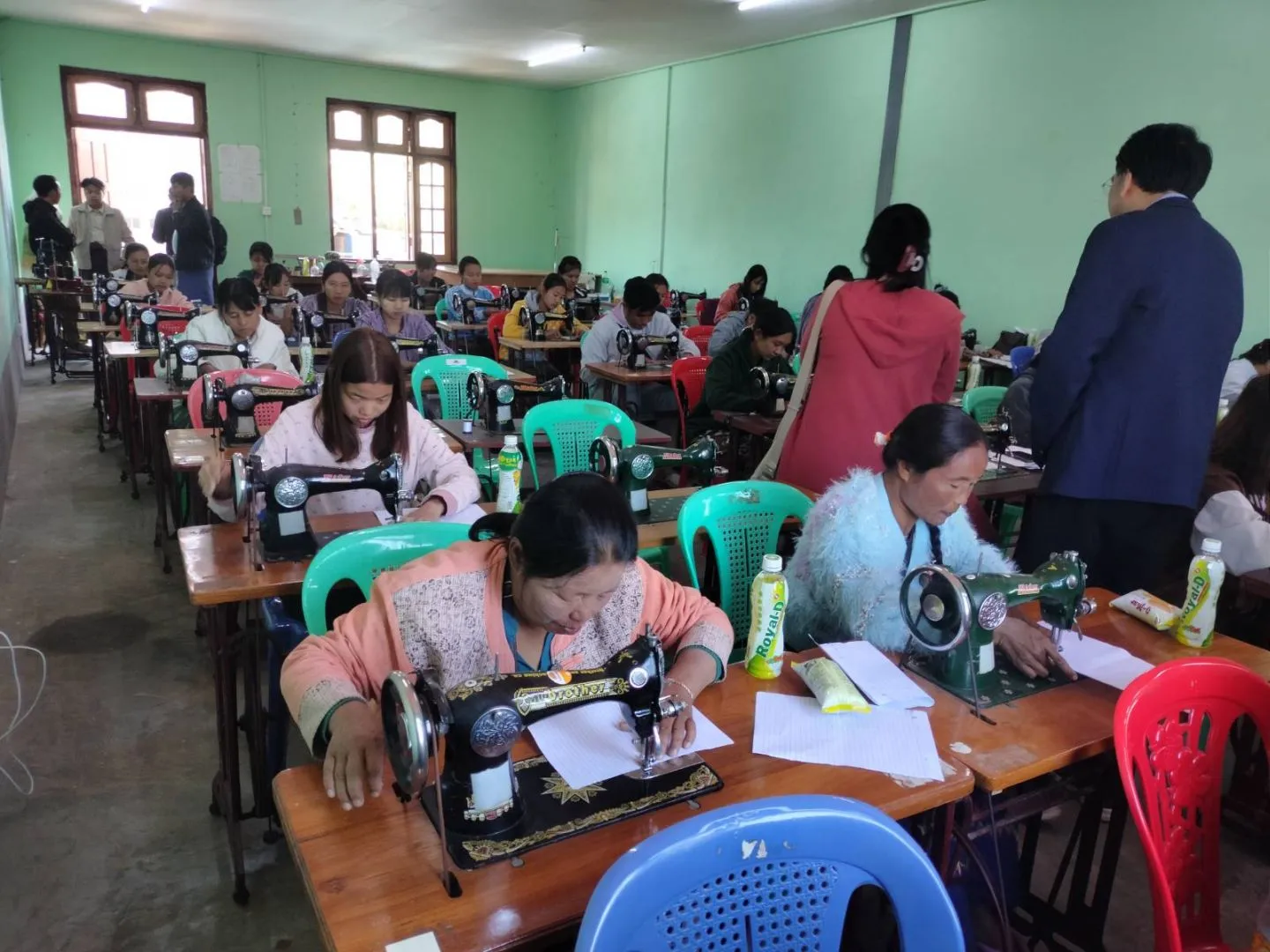 Women are practising with sewing machines in the training and a woman & a man are checking them.